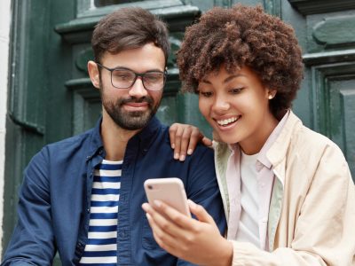 Attractive hipster and his dark skinned girlfriend watch funny video online, make shopping, use electronic gadgets, pose against ancient building door background. People, relationship, youth concept